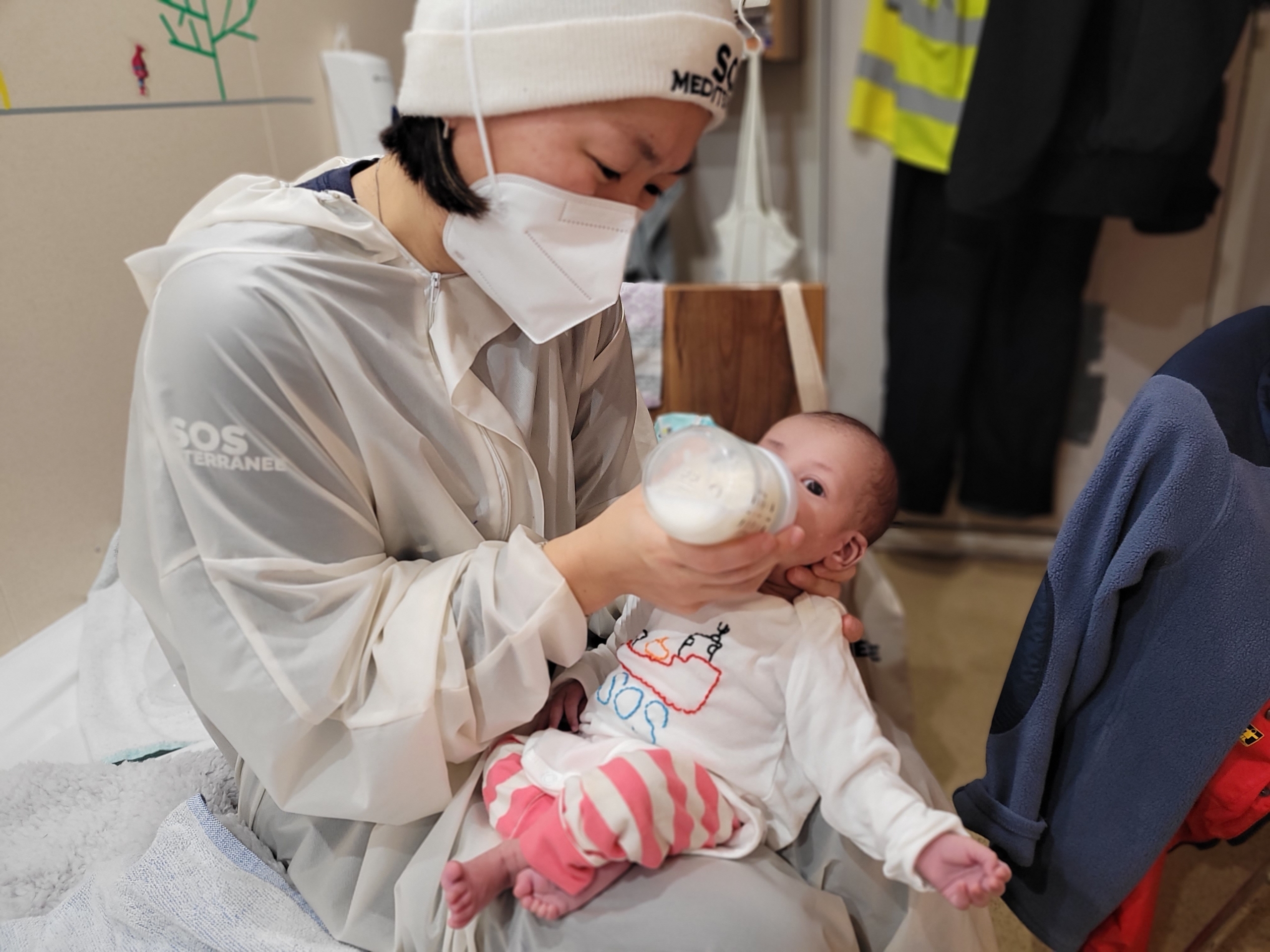 An Asian woman wearing a white face mask, white gown, and white hat giving a bottle to a white baby.